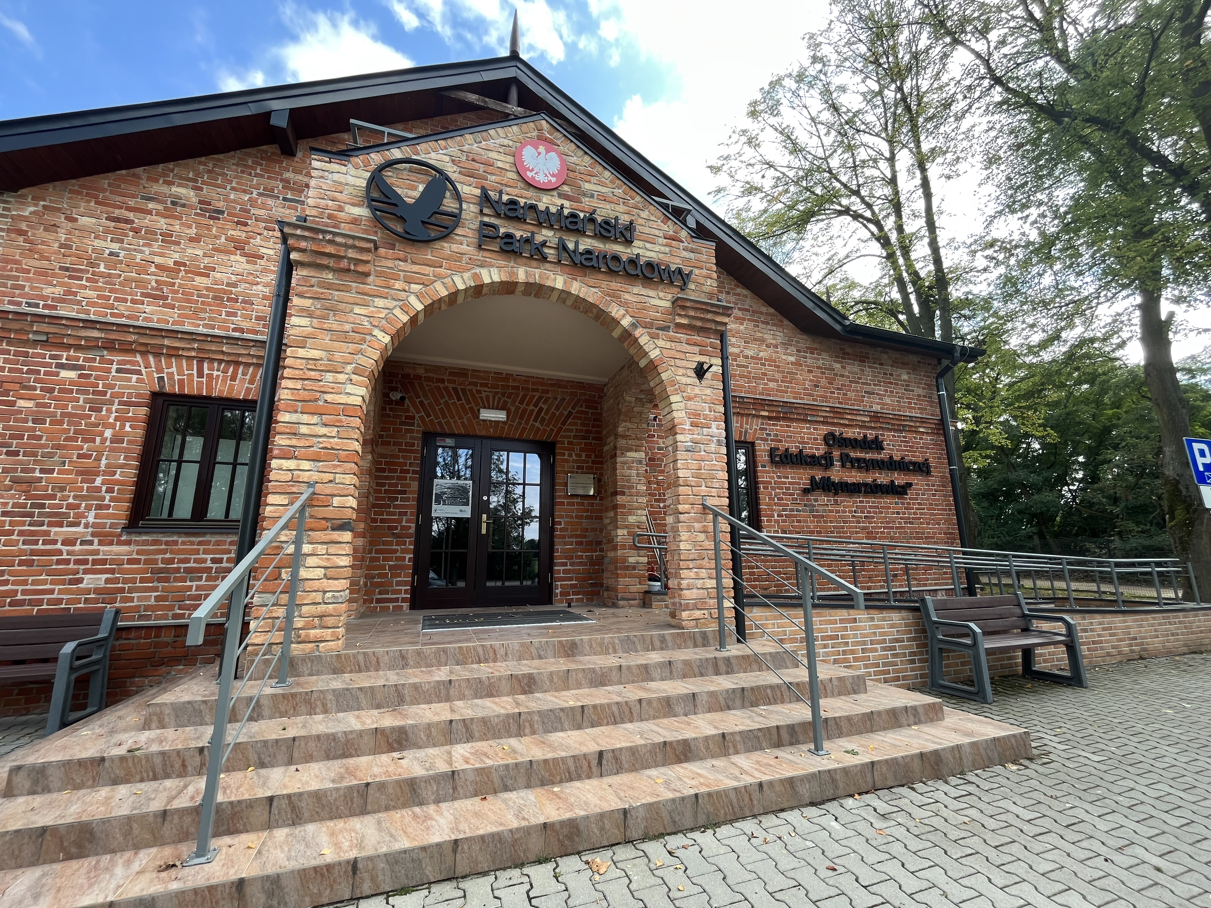 Wejście do Ośrodka Edukacji Przyrodniczej „Młynarzówka” w Kurowie. Na wprost schody z płytek i fasada budynku Ośrodka Edukacji Przyrodniczej "Młynarzówka" Narwiańskiego PN zrobiona z czerwonej cegły. Z prawej strony podjazd na wózków. Po obu stronach schodów ławki. W tle drzewa.