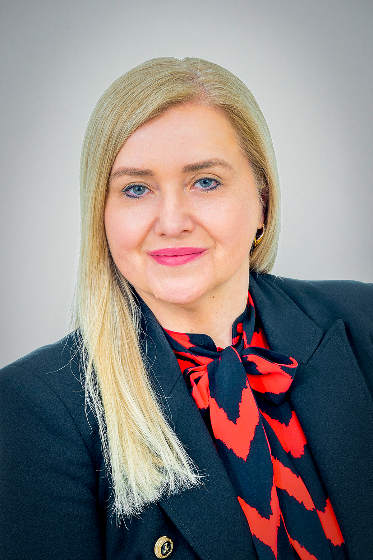 Widoczna górna część sylwetki kobiety z pogodnym wyrazem twarzy. Włosy jasny blond, długie proste, rozpuszczone. Kobieta zwrócona przodem do kamery. Ma na sobie granatowy żakiet, pod nim bluzkę w kolorze granatowo-pomarańczowym.