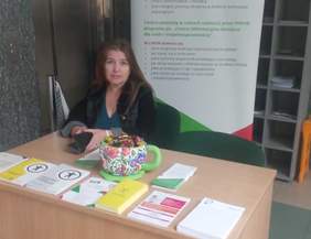 Pokaż zdjęcie: Przy biurku siedzi kobieta z długimi włosami. Na biurku leżą ulotki, za plecami stoi rollup z logo PFRON.