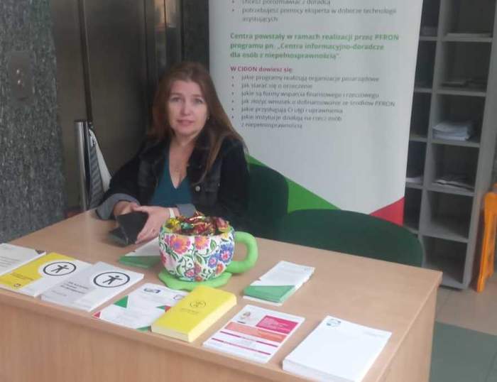 Pokaż zdjęcie: Przy biurku siedzi kobieta z długimi włosami. Na biurku leżą ulotki, za plecami stoi rollup z logo PFRON.