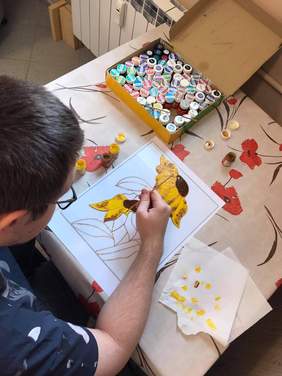 Pokaż zdjęcie: Widok mężczyzny pochylonego nad kartką papieru, trzymającego w prawej dłoni pędzel i malującego żółte kwiaty, przed nim na stole ustawione są kolorowe słoiczki z farbami.