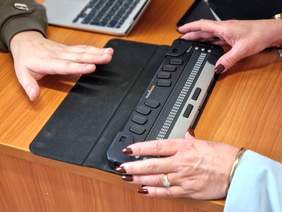 Pokaż zdjęcie: Na stole znajduje się sprzęt specjalistyczny dla osób niewidomych. Widoczne są dłonie dwóch osób, które dotykają sprzęt. 