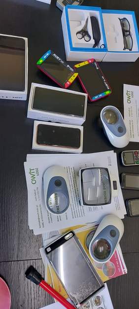 Pokaż zdjęcie: Na stole ułożone są różnego rodzaju sprzęty i technologie wspomagające osoby z niepełnosprawnościami. 