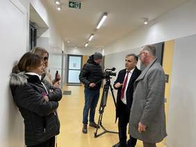 Pokaż zdjęcie: Dwóch mężczyzn rozmawia z dwiema kobietami w holu w bloku. Pierwszy z mężczyzna ubrany jest w szary płaszcz drugi mężczyzna ubrany jest w czarny płaszcz białą koszulę oraz czerwony krawat. Za nimi stoi mężczyzna ubrany puchową kurtkę i sza