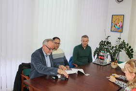 Pokaż zdjęcie: Zdjęcie trzech mężczyzn siedzących przy stole, pierwszy od lewej starszy mężczyzna poodpisuje umowę. Na stole stoją pieczątki.