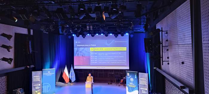 Pokaż zdjęcie: Widok na salę konferencyjną, która jest zaciemniona, widać także prelegenta ubranego w garnitur, na tle prezentacji