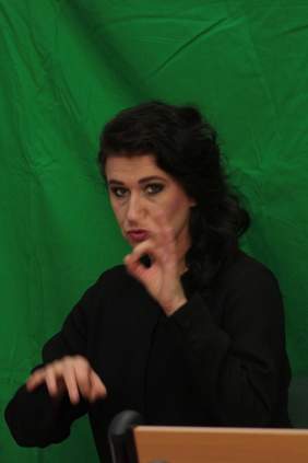 Pokaż zdjęcie: Kobieta siedzi na tle zielonej ściany używa języka migowego