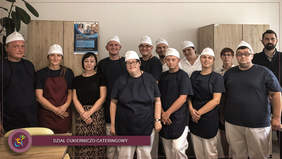 Pokaż zdjęcie: W pomieszczeniu w 2 rzędach stoi 13 osób w większości ubranych w czarne koszulki i białe spodnie na które mają założone fartuchy, na głowach mają białe czapki kucharskie.