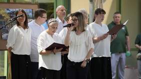 Pokaż zdjęcie: Młoda kobieta śpiewa do mikrofonu. W tle widać chór. Wszyscy występujący są ubrani na galowo. 