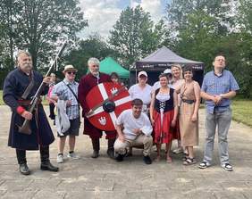 Pokaż zdjęcie: Grupa osób pozuje do wspólnego zdjęcia, wśród nich są rekonstruktorzy w historycznych strojach – jeden trzyma długą strzelbę, drugi tarczę z orłem – oraz uczestnicy i pracownicy warsztatu, niektórzy w przebraniach tematycznych. 