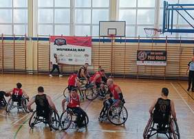 Pokaż zdjęcie: Zdjęcie przedstawia mecz koszykówki na wózkach w hali sportowej. Zawodnicy w koszulkach czerwonych  i czarnych  rywalizują na boisku. W tle widoczne są banery z nazwami klubów oraz osoby obserwujące mecz. Drewniana podłoga hali oznaczona jest liniami boiska do koszykówki, a kosze znajdują się po obu stronach sali.