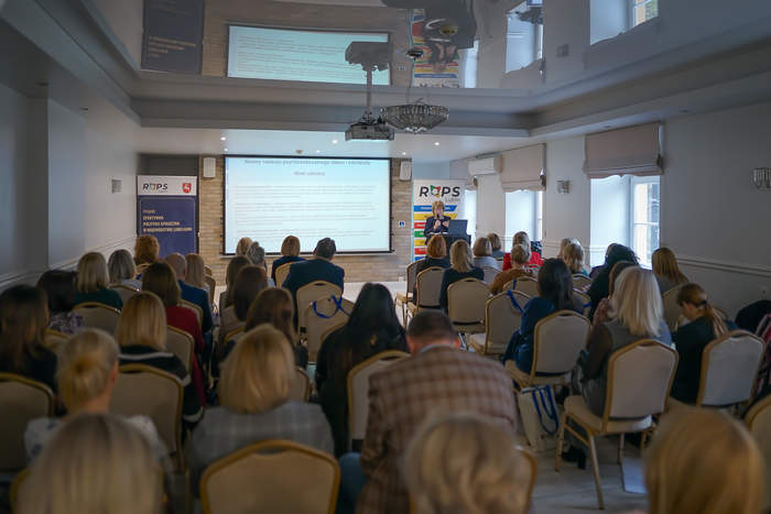 Pokaż zdjęcie: Zdjęcie z tyłu sali konferencyjnej w kierunku prelegenta, cała sala wypełniona jest uczestnikami, na ścianie slajd z prezentacji.