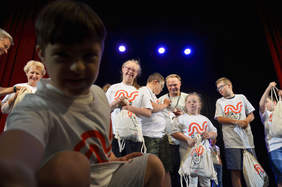 Pokaż zdjęcie: Na scenie uśmiechnięte dzieci ubrane w koszulki z napisem Nieprzetarty  Szlak w tle opiekunowie