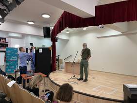 Pokaż zdjęcie: Na scenie stoi mężczyzna z siwą brodą w okularach ubrany w spodnie bojówki i jednolitą koszulę z kieszeniami na piersiach. Na widowni siedzą ludzie, operator poprawia statyw z kamerą.