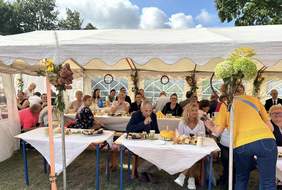 Pokaż zdjęcie: Grupa kilkunastu osób siedzi przy kliku stolikach ustawionych pod dużym namiotem. Stoły przykryte są białymi obrusami. Ustawione są na nich różne napoje oraz słodki poczęstunek. W tle widoczne lekko zachmurzone niebo oraz drzewa liściaste.