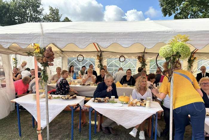 Pokaż zdjęcie: Grupa kilkunastu osób siedzi przy kliku stolikach ustawionych pod dużym namiotem. Stoły przykryte są białymi obrusami. Ustawione są na nich różne napoje oraz słodki poczęstunek. W tle widoczne lekko zachmurzone niebo oraz drzewa liściaste.