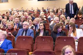 Pokaż zdjęcie: Zdjęcie przedstawia widownię w teatrze lub sali konferencyjnej, wypełnioną dorosłymi osobami siedzącymi w rzędach bordowych foteli. Wiele osób jest uśmiechniętych i patrzy w kierunku sceny (poza kadrem). Na pierwszym planie widoczny jest rząd foteli, oznaczony numerami (np. 6, 7, 8). Po prawej stronie, na górze, mężczyzna w ciemnym garniturze i krawacie stoi przy rzędzie i wchodzi na widownię.