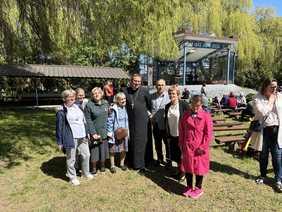 Pokaż zdjęcie: Pośrodku zdjęcia stoi mężczyzna ubrany czarną sutannę obok mężczyzna wiosna i marynarce i Kobiety elegancko ubrane. Wszyscy się uśmiechają mrużąc oczy od rażącego słońca W tle scena nad którą jest napis wierzę w syna Bożego. 