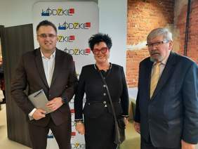 Pokaż zdjęcie: Do zdjęcia pozują trzy osoby: kobieta i dwóch mężczyzn. Uśmechają się. Są elegancko i szykownie ubrani. 