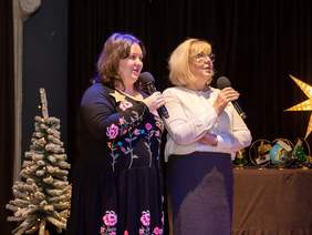 Pokaż zdjęcie: Scena, na środku dwie kobiety, śpiewają do mikrofonu, patrzą na ekran z tekstem kolędy.