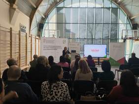 Pokaż zdjęcie: Zdjęcie z tyłu sali w kierunku prelegenta, uczestnicy spotkania siedzą na krzesełkach.