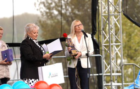 Scena plenerowa, na której stoją dwie kobiety. Kobieta po prawej, w białej marynarce i okularach, przemawia do mikrofonu, trzymając w ręku tę samą statuetkę co na pierwszym zdjęciu oraz czerwoną różę. Kobieta po lewej, w ciemnej marynarce, trzyma białą torbę prezentową z logo „Pomorze Zachodnie” i przegląda dokumenty. U dołu kadru widoczne są kolorowe balony dekorujące przód sceny.