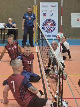 Grupa zawodników i organizatorów stoi na hali sportowej przed banerami PFRON, Ministerstwa Sportu i Turystyki oraz Klubu Sportowego START Szczecin. Na środku znajduje się kolorowa maskotka w stroju sportowym.