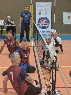 Grupa zawodników i organizatorów stoi na hali sportowej przed banerami PFRON, Ministerstwa Sportu i Turystyki oraz Klubu Sportowego START Szczecin. Na środku znajduje się kolorowa maskotka w stroju sportowym.