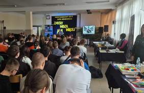 Pokaż zdjęcie: Zdjęcie z tyłu sali w kierunku sceny podczas wydarzenia. Z boku sali wzdłuż okien ustawiono stoiska informacyjne.