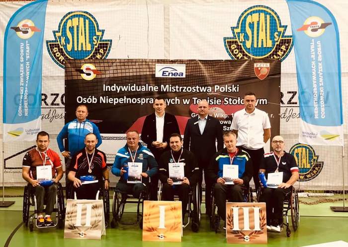 Pokaż zdjęcie: Zawodnicy na wózkach inwalidzkich z medalami na tle podium i ścianki promocyjnej Mistrzostw