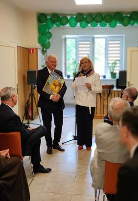 Pokaż zdjęcie: Na środku elegancko ubrani, usmiechnięci kobieta i mężczyzna. Kobieta przekazauje mężczyżnie obraz