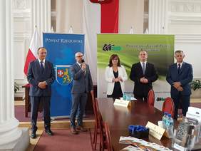 W szeregu stoi pięć osób czterech mężczyzn i w środku kobieta. Przestronna sala urzędu. Mężczyzna przemawia do mikrofonu.