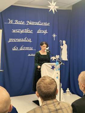 Pokaż zdjęcie: Przy mównicy stoi kobieta w czarneju sukience, w tle granatowa kotara z hasłem: W Boże Narodzenie wszystkie drogi prowadzą do domu.