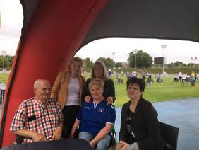 Pokaż zdjęcie: Na zewnątrz pod namiotem, przy stoliku siedzą cztery osobę, za nimi stoi kobieta i mężczyzna