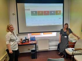 Pokaż zdjęcie: Po dwóch stronach tablicy multimedialnej stoją dwie panie. Na tablicy wyświetlone są zagadnienia zwiazane z Systemem Obsługi Wsparcia. Panie się uśmiechają. 