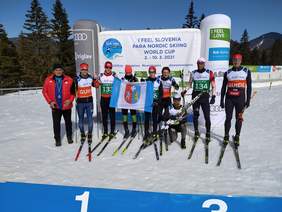 9 osób, trener i 8 narciarzy na tle baneru mistrzostw trzymają flagę woj. podkarpackiego, słoneczny dzień