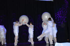 Pokaż zdjęcie: Na scenie grupa dzieci ubranych na biało tańczy z dużymi białymi piłkami