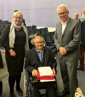 Pokaż zdjęcie: Trzy uśmiechnięte osoby, kobieta i dwóch mężczyzn. Mężczyzna na środku siedzi na wózku inwalidzkim. W rękach trzyma pamiątkowy dyplom