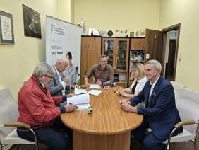 Pokaż zdjęcie: Przy owalnym stole siedzi pięć osób: kobieta i czterech mężczyzn. Podpisują umowę. Na stole leżą różne dokumenty, pieczątki i artykuły biurowe. 