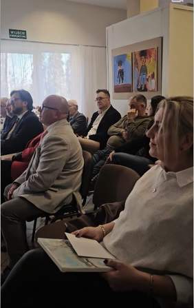 Pokaż zdjęcie: Na krzesłach siedzą ludzie - uczestnicy dorocznej wystawy prac plastycznych. Na ścianie wiszą dwa obrazy. W tle widać także okno. 