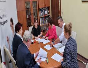 Pokaż zdjęcie: pomieszczenie Oddziału po środku owalny stół przy którem siedzą cztery osoby na stole dokumenty które podpisują upoważnione osoby 