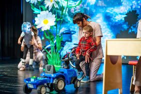 Kadr ze sceny teatralnej lub terapeutycznej z udziałem dzieci i opiekunów. Na pierwszym planie po prawej stronie kobieta w jasnym stroju medycznym klęczy, asekurując stojącego chłopca w czerwonej koszuli w kratę. W tle, po lewej stronie, druga opiekunka wspiera dziewczynkę korzystającą z ortez pionizujących. Wokół znajdują się rekwizyty: niebieski zabawkowy traktor na pierwszym planie oraz gigantyczne, sztuczne stokrotki w tle. Tłem sceny jest niebieska, świetlna projekcja.
