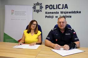Pokaż zdjęcie: Przedstawiciel PFRON i funkcjonariusz Policji podpisują dokumenty przy stole w siedzibie Komendy Wojewódzkiej Policji w Lublinie, na tle logotypów instytucji.