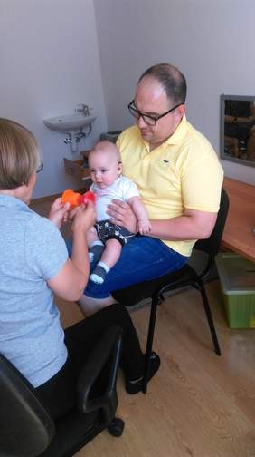 Pokaż zdjęcie: Kobieta i mężczyzna siedzący naprzeciwko siebie na krzesłach. Mężczyzna trzyma małego chłopca na kolanach a kobieta pokazuje kolorowe kubeczki.