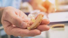 Pokaż zdjęcie: Na otwartej dłoni leży aparat słuchowy. Tło zdjęcia jest rozmyte. 