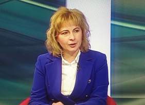 Pokaż zdjęcie: Przy szklanym stole siedzi kobieta, zdjęcie robione z bliska. Kobieta blondynka ubrana w niebieski garnitur i białą koszulę