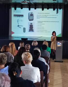 Pokaż zdjęcie: Zdjęcie zrobione z tyłu sali podczas konferencji, przy mikrofonie kobieta w brązowej sukience. W tle slajd z prezentacji.