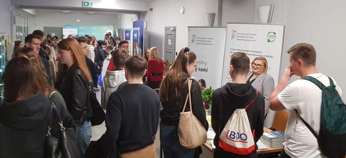 Pokaż zdjęcie: Trzy młode osoby stoją przy stoisku Oddziału Wielkopolskiego PFRON, przy którym obecna jest kobieta udzielająca informacji. W tle widoczne są banery: PFRON i CIDON. Na korytarzu znajduje się duża grupa osób, która odwiedza stoiska innych wystawców.