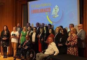 Pokaż zdjęcie: W sali znajduje się grupa kilkunastu osób, która stoi na tle dużego ekranu. Wśród nich są dwie poruszające się na wózku. Na ekranie wyświetlony jest napis: „XIX edycja Konkursu Lodołamacze”, jak również widoczne jest zdjęcie statuetki „lodołamacza”.