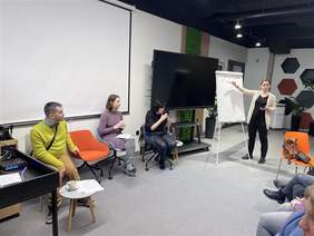 Pokaż zdjęcie: Na zdjęciu widać cztery osoby biorące udział w warsztatach w nowoczesnej sali. Jedna z nich stoi przy flipcharcie i zapisuje informacje, podczas gdy pozostałe osoby siedzą na krzesłach, słuchając i dyskutując.